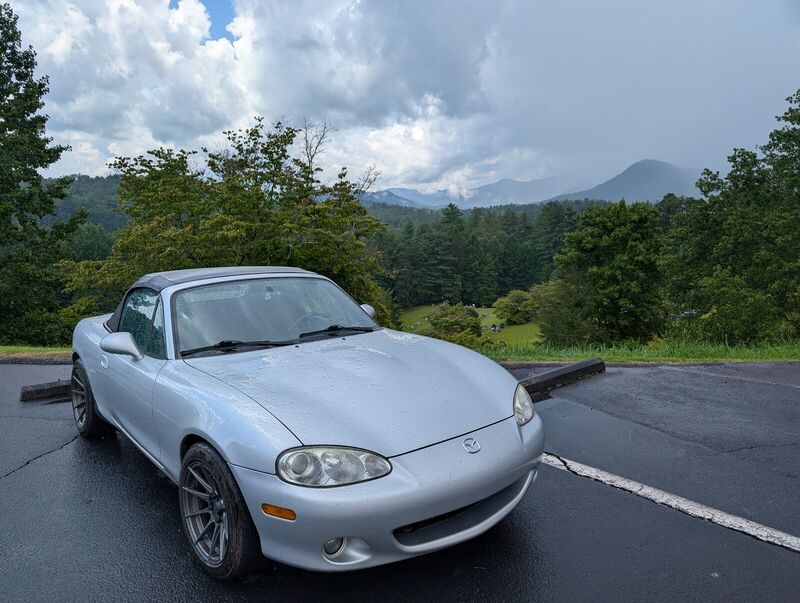 The Miata parked at a mountain overlook on wet pavement after rain, top up, ridgelines behind it.