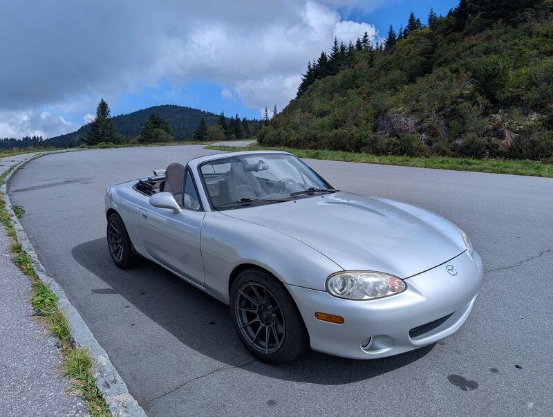 The Miata parked on the shoulder of a mountain road with the top down, forested ridgelines behind it.