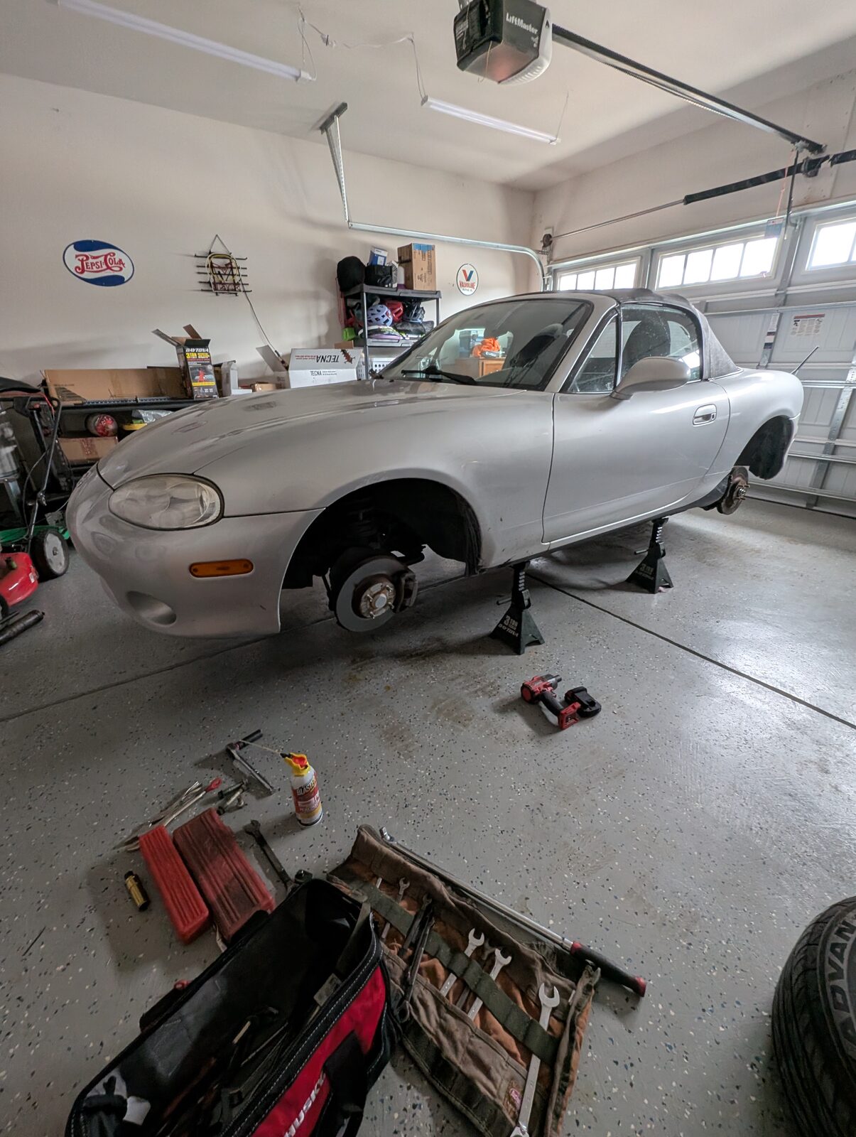 The Miata up on jack stands in a garage with the front wheel removed and tools spread across the floor during the coilover installation.