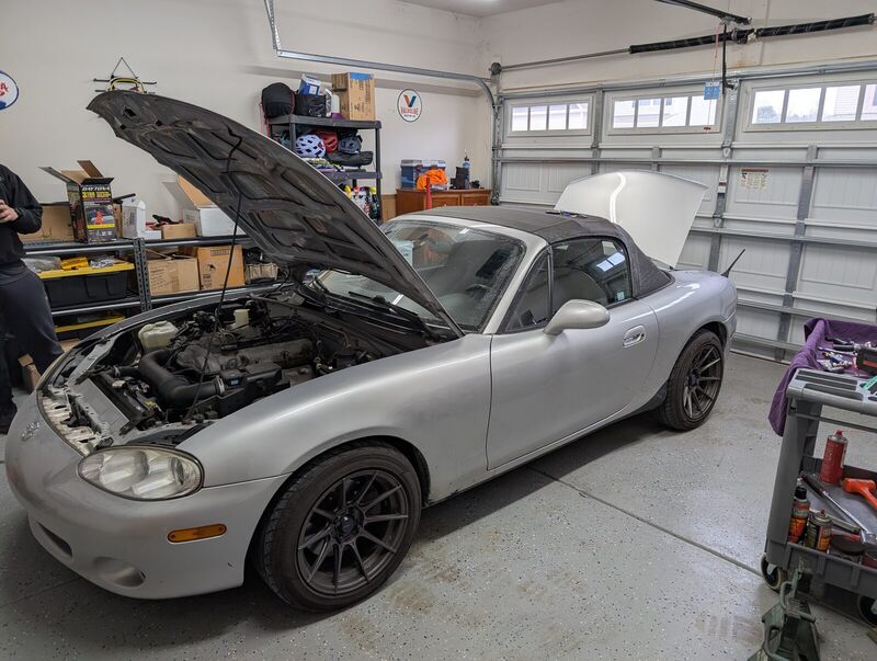 The Miata back on its wheels in a garage with the hood up and a tool cart alongside.