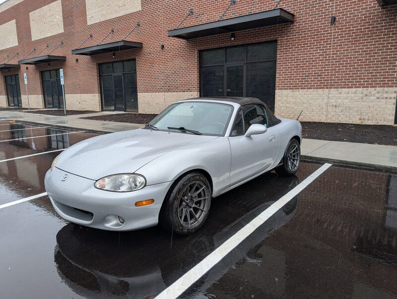 The Miata on wet pavement outside a brick building, top up, on its gunmetal wheels.