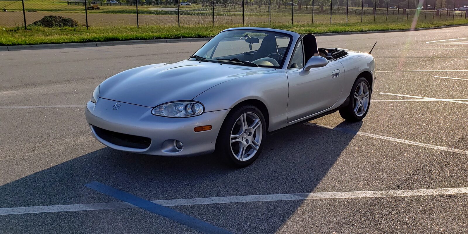 The silver Miata parked in an empty lot in low evening sun on the day it was bought, still on its original wheels with the top down.