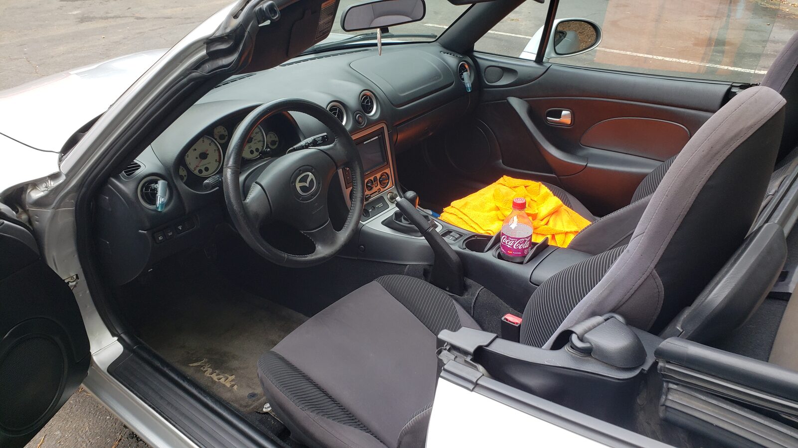 The Miata&rsquo;s interior seen through the open driver&rsquo;s door, showing the stock steering wheel, gauges, seats and the original shift knob.