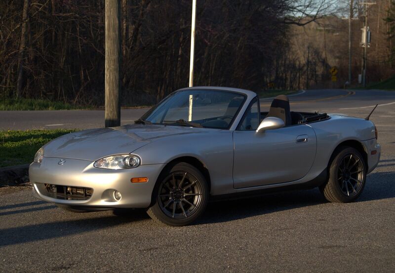 Three-quarter front view of the Miata in golden evening light at the side of a country road, on the day the new gunmetal wheels were fitted.