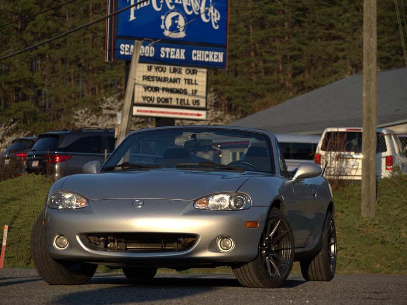 The Miata facing the camera in evening light, parked beneath a roadside restaurant sign.